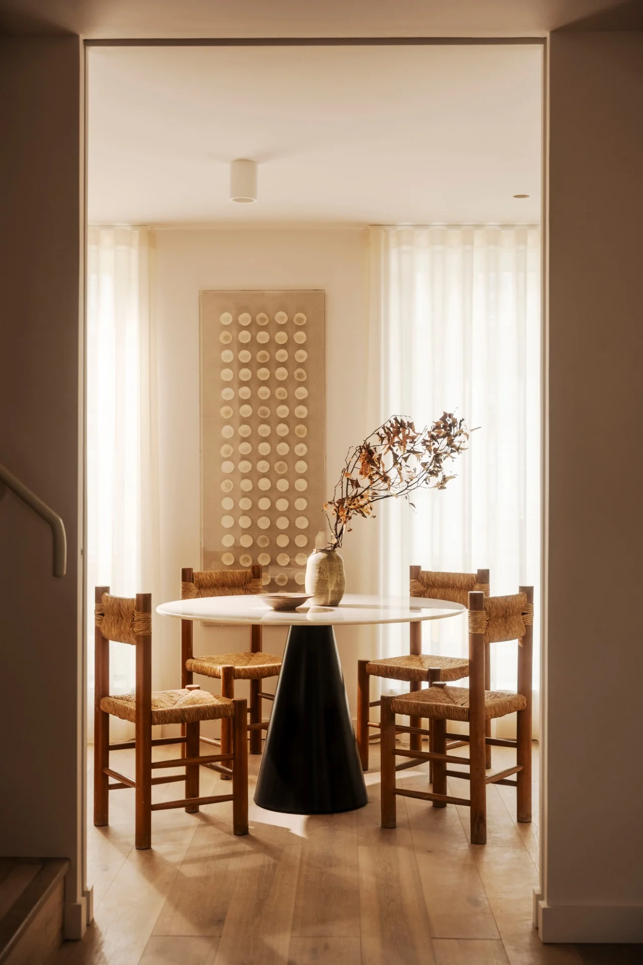 Dining table with rush-seat chairs beneath a textile wall hanging, framed by a doorway