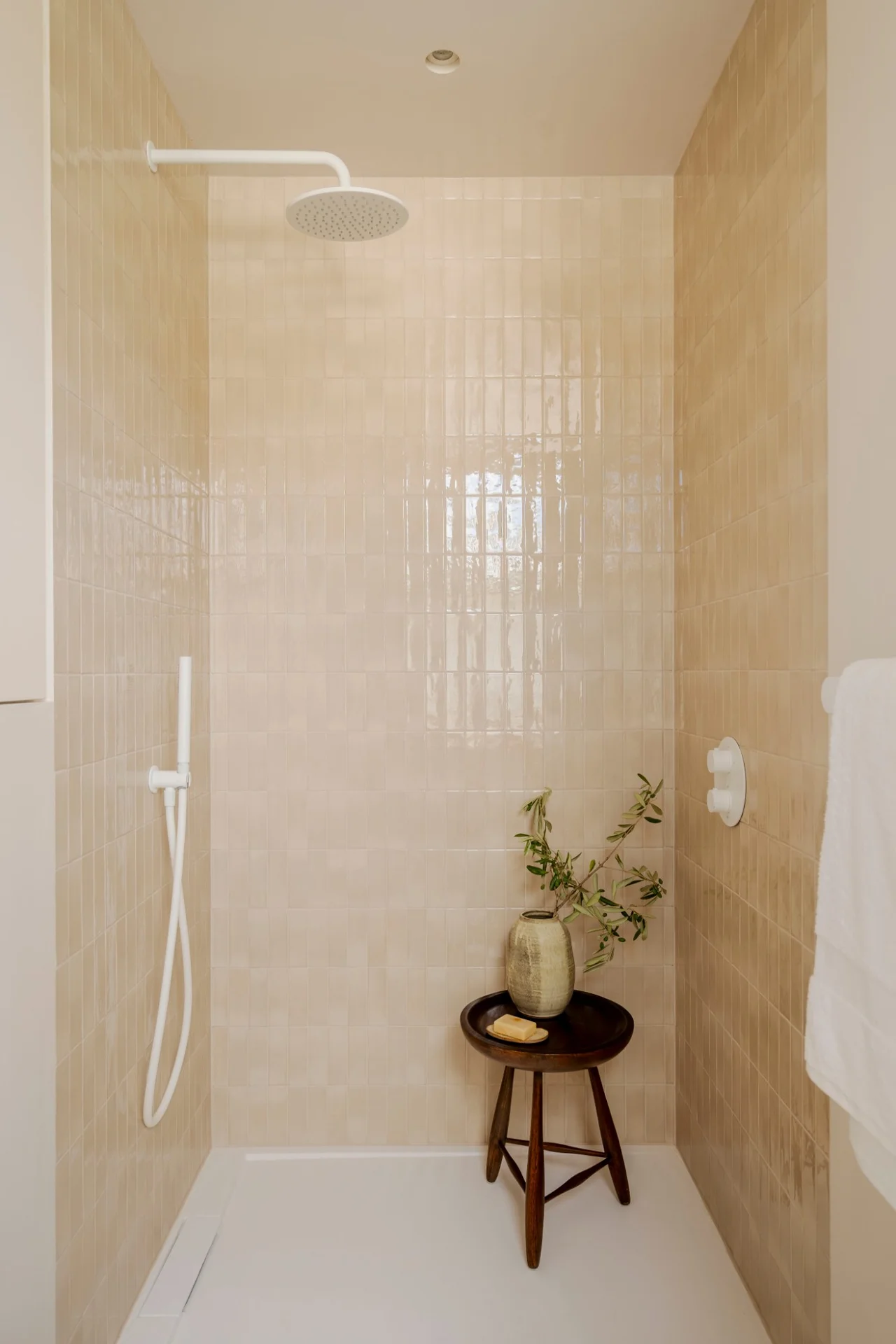 Shower lined in glazed cream tiles, with a wooden stool and olive branch